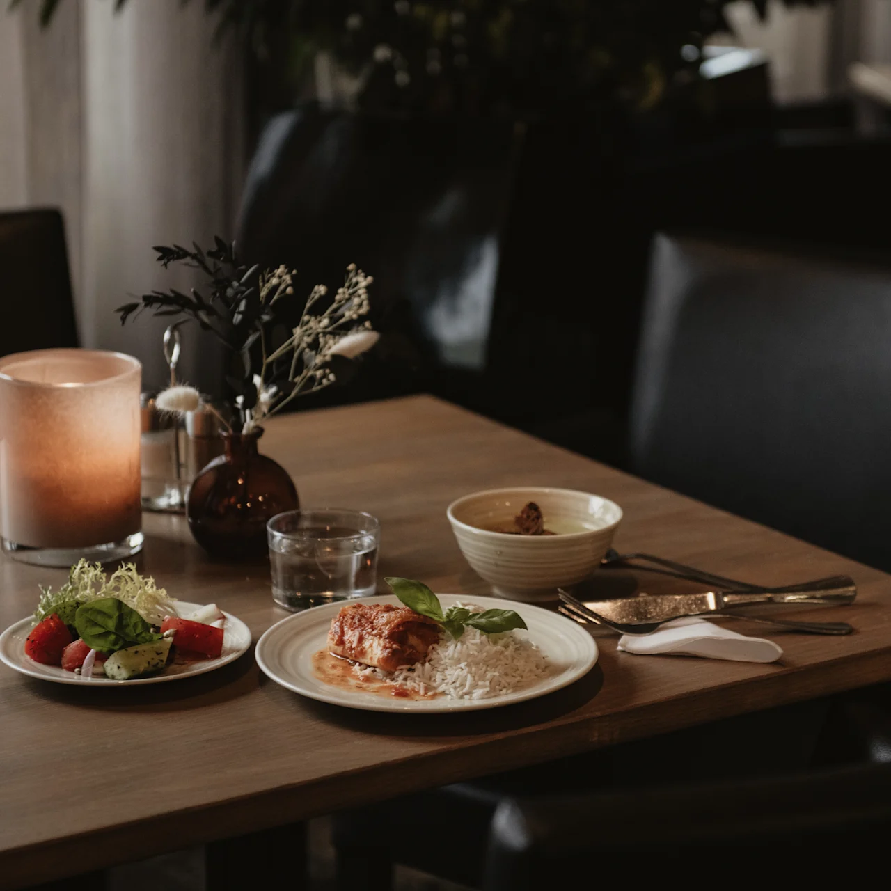 A wooden table holds a plated meal with salad, rice, and meat, accompanied by a candle and a bowl, in a dimly lit dining area.