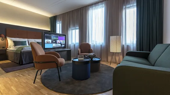 A modern hotel room with a double bed, television displaying "Quality Hotel," seating area, and floor lamp, surrounded by large windows with sheer curtains.