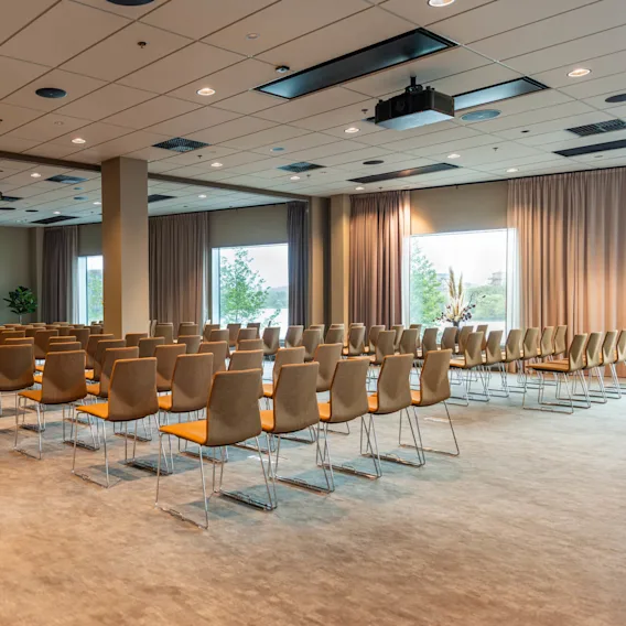 Rader med tomme stoler vender mot en stor prosjektorskjerm som viser et bylandskap, i et romslig konferanserom med store vinduer og beige gardiner.