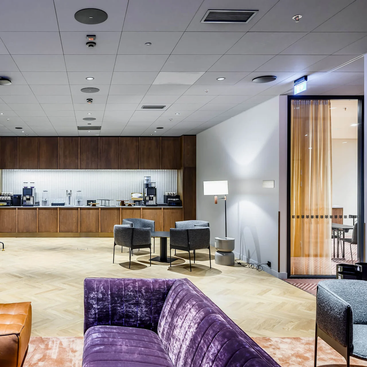 Coffee station featuring machines and cups is set on a wooden counter in a modern lounge area with gray chairs, purple sofa, and leather ottoman on a light wood floor.