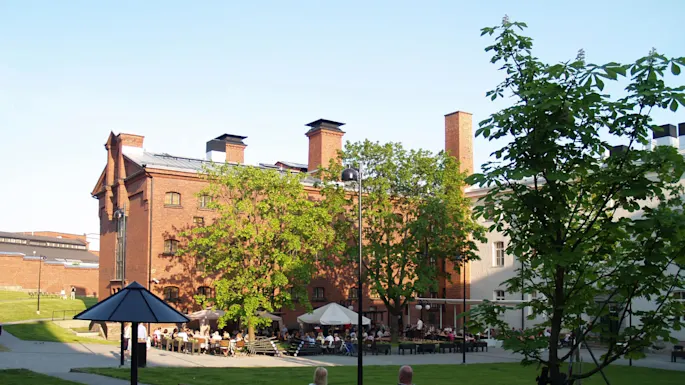 Mennesker slapper av ved utendørs bord i nærheten av en mursteinsbygning med grøntområder, i en rolig, urban parkinnstilling.