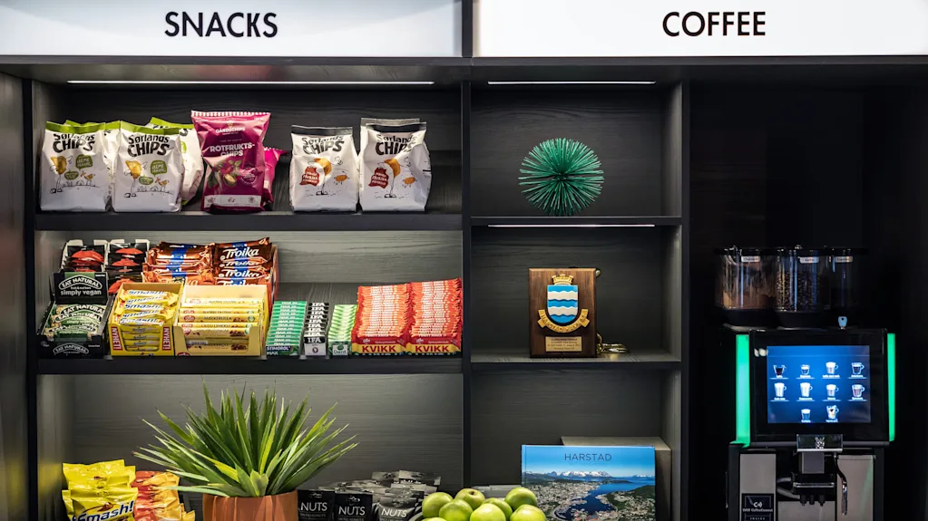Snacks och kaffestation med olika chips, choklad och nötter på hyllor. En kaffemaskin, gröna äpplen i en skål och en liten växt pryder utrymmet. Text: "SNACKS," "COFFEE," "HARSTAD."