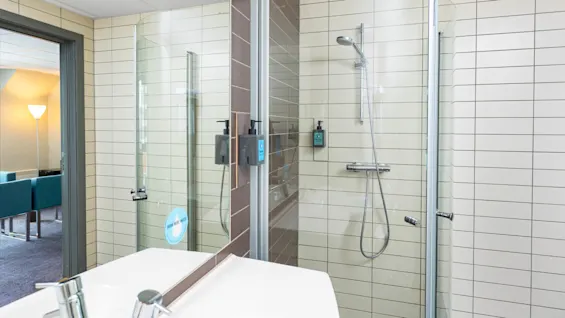 A modern shower with glass door and wall-mounted soap dispensers stands in a tiled bathroom. Through a doorway, an adjacent room with a blue sofa and floor lamp is visible.