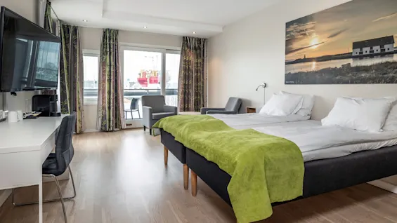 Ett prydligt hotellrum med en dubbelsäng täckt med en grön pläd, vänd mot en TV och ett skrivbord; stora fönster med mönstrade gardiner erbjuder utsikt över en hamn.
