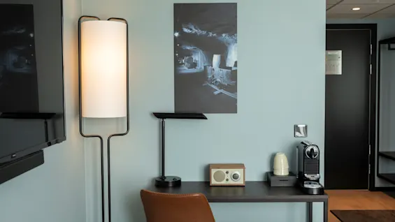 Et koselig hotellrom med et skrivebord, kaffemaskin, radio, en stilig gulvlampe og et unikt kunstverk med gruvetema.