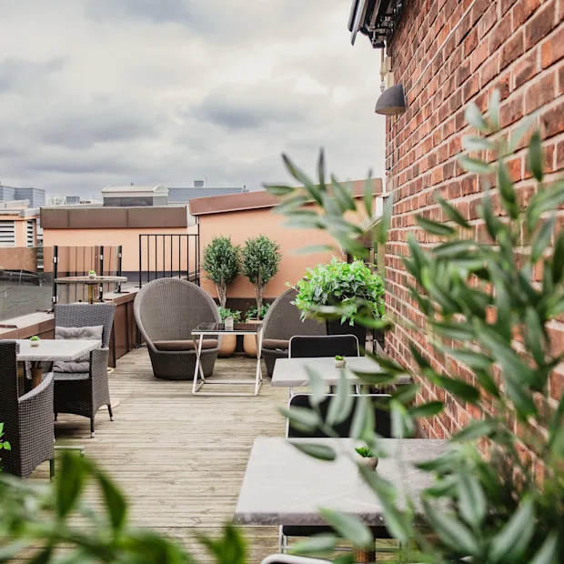 A rooftop terrace with outdoor seating, lush green plants, and a brick wall under a cloudy sky.