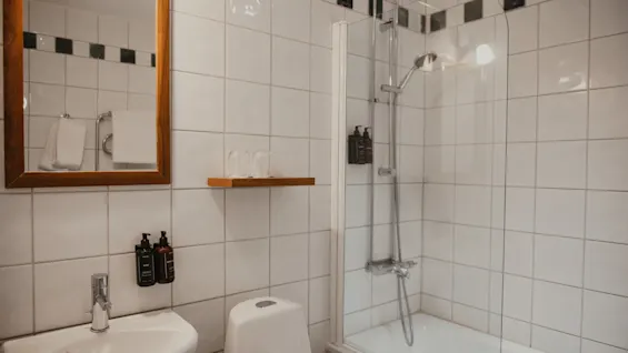 Et lyst og funktionelt hotelbadeværelse med hvide flisevægge, en vask, toilet og en bruser-badekar-kombination.