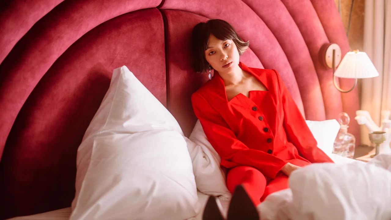 A person in a red outfit sits on a bed against a plush, curved headboard. White pillows surround them. A bedside lamp glows warmly beside the bed.