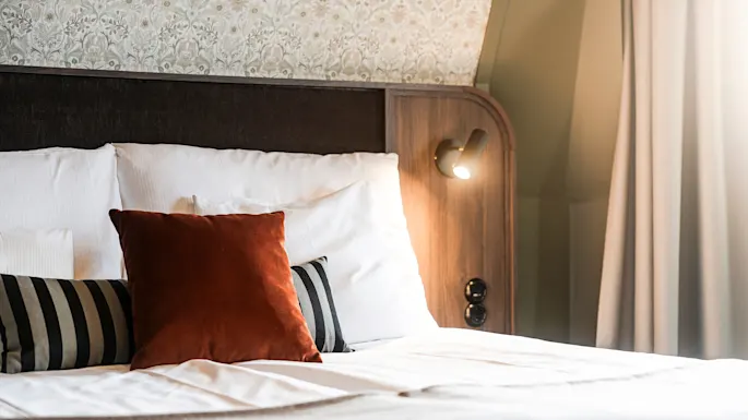 En pent oppredd seng med puter, én brun, mot et elegant sengegavl, med en tent veggmontert lampe.