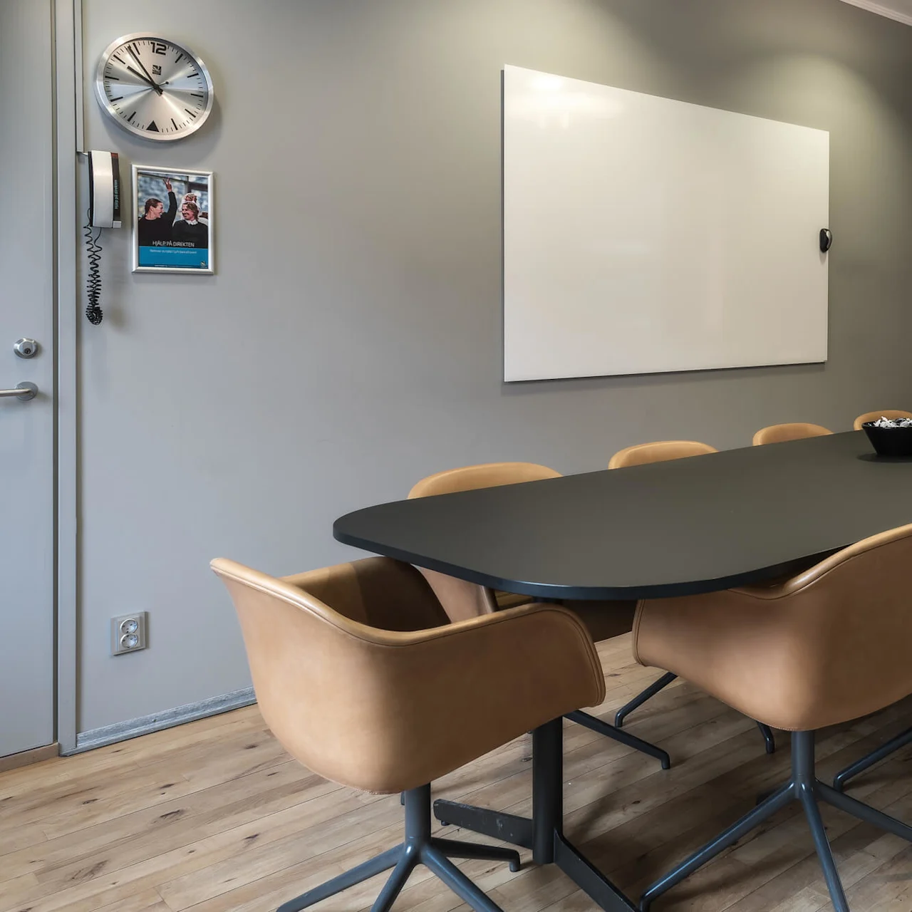 A conference table with brown chairs surrounds a black table with a candy bowl. Walls feature a clock, a whiteboard, and a mounted screen. A wall poster reads "WE PUT DIRECTORS".