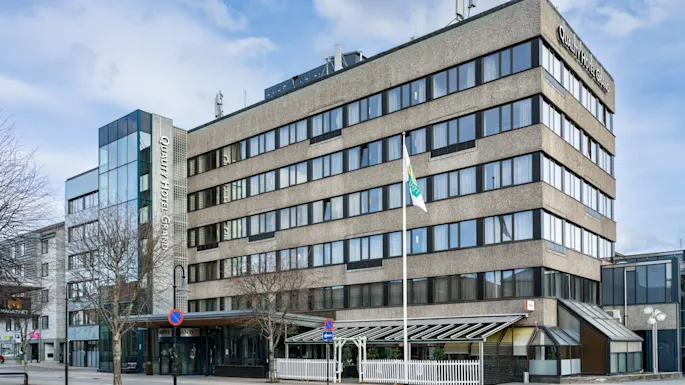 A multi-story hotel building stands quietly on a city street; its sign reads "Quality Hotel Maritim," with flags fluttering nearby under a blue sky.