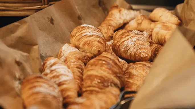 Croissanterna er stablet, drysset med melis, på brunt papir. De ligger i en flettet kurv og skaper en koselig bakeriatmosfære.