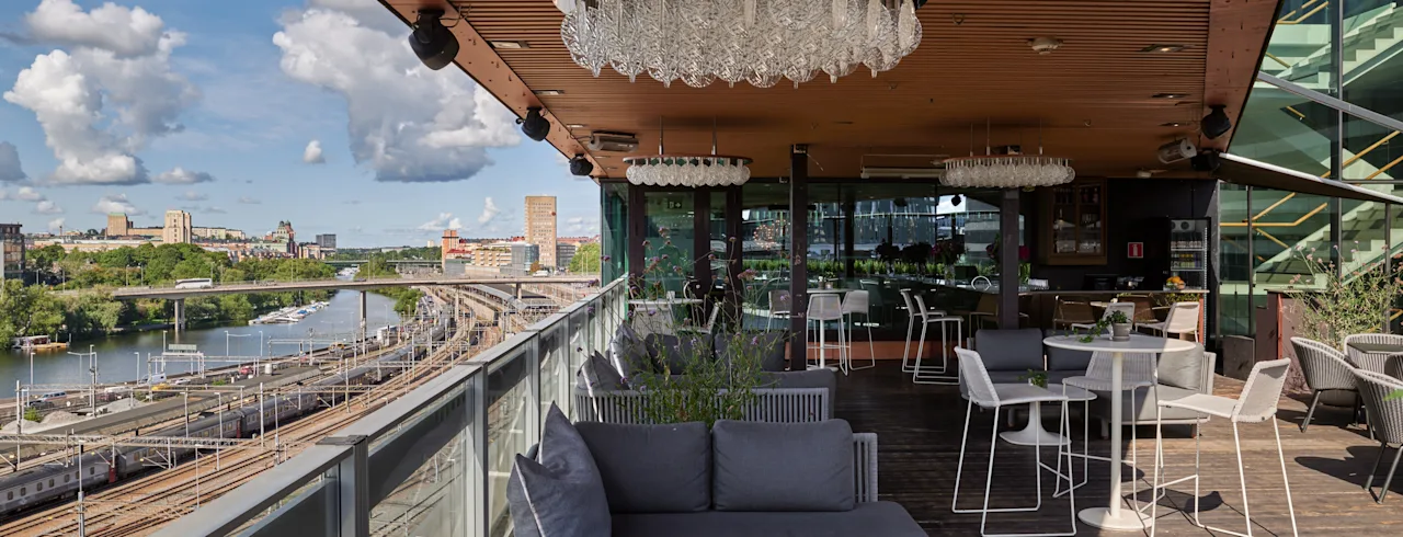 Utomhusterrass med moderna möbler och ljuskronor; har utsikt över järnvägsspår och en flod. Omgiven av stadssilhuett under en delvis molnig himmel.
