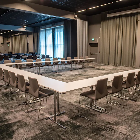U-format konferensbord omgivet av stolar står i ett rymligt, mattbelagt mötesrum med gardinbeklädda fönster och mjuk belysning.