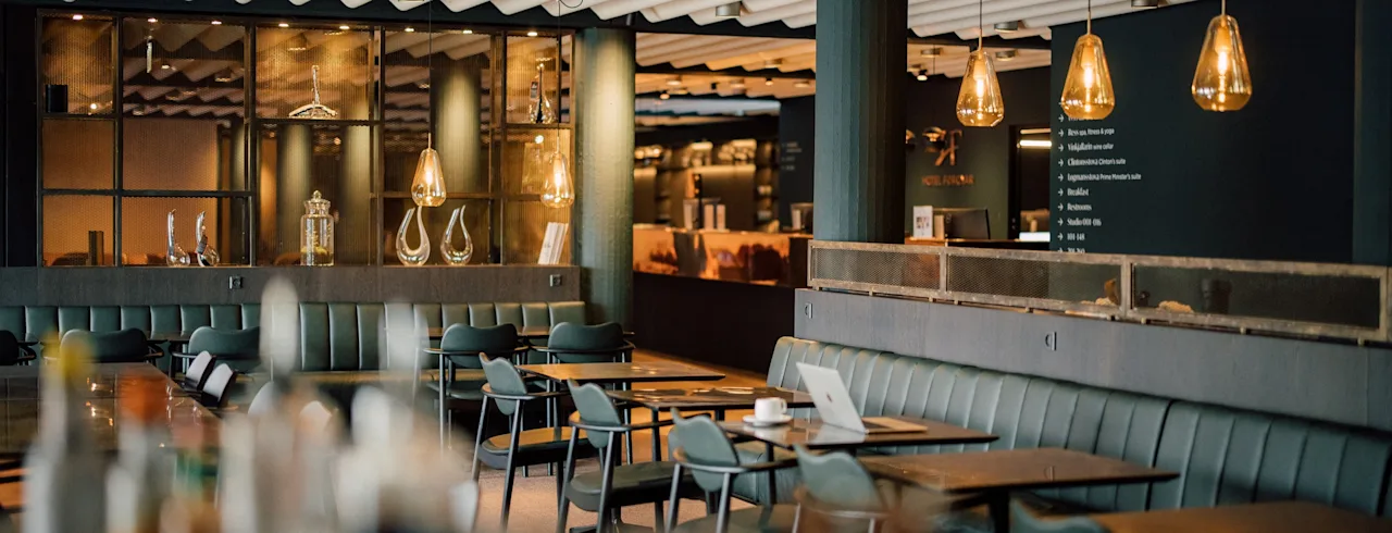 A modern restaurant features green velvet seating and wooden tables. Warm pendant lights create an inviting atmosphere. A laptop and cup are on one table.
