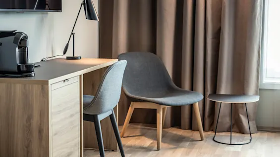 En moderne indretning med et træbord med kaffemaskine og lampe. To grå stole og et lille sidebord står ved brune gardiner.