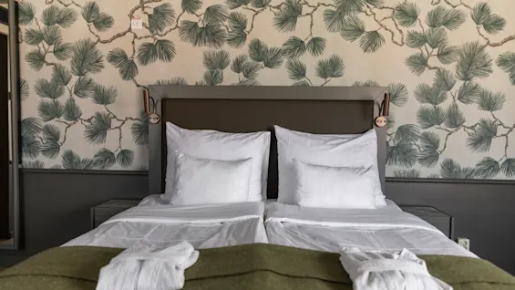 Viihtyisä hotellihuoneen sänky valkoisilla liinavaatteilla, vihreällä peitolla ja kylpytakeilla, luontoaiheista tapettia vasten.