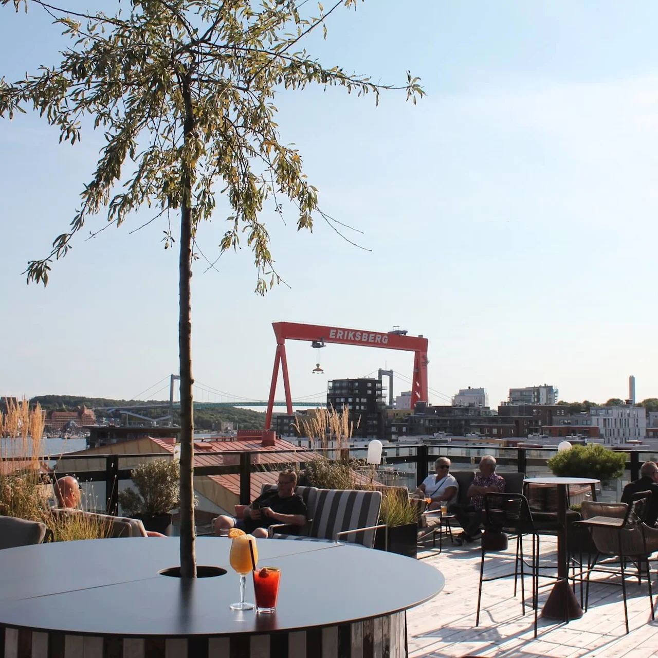 Table with drinks stands in a rooftop bar, surrounded by people seated and socializing; in the background, the Eriksberg crane and cityscape are visible.