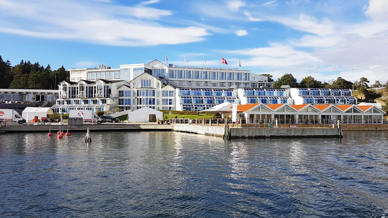 Large waterfront building displaying Danish flag, surrounded by woods and a calm sea. Nearby, small orange-roofed structures align the shoreline; a clear sky enhances the peaceful setting.