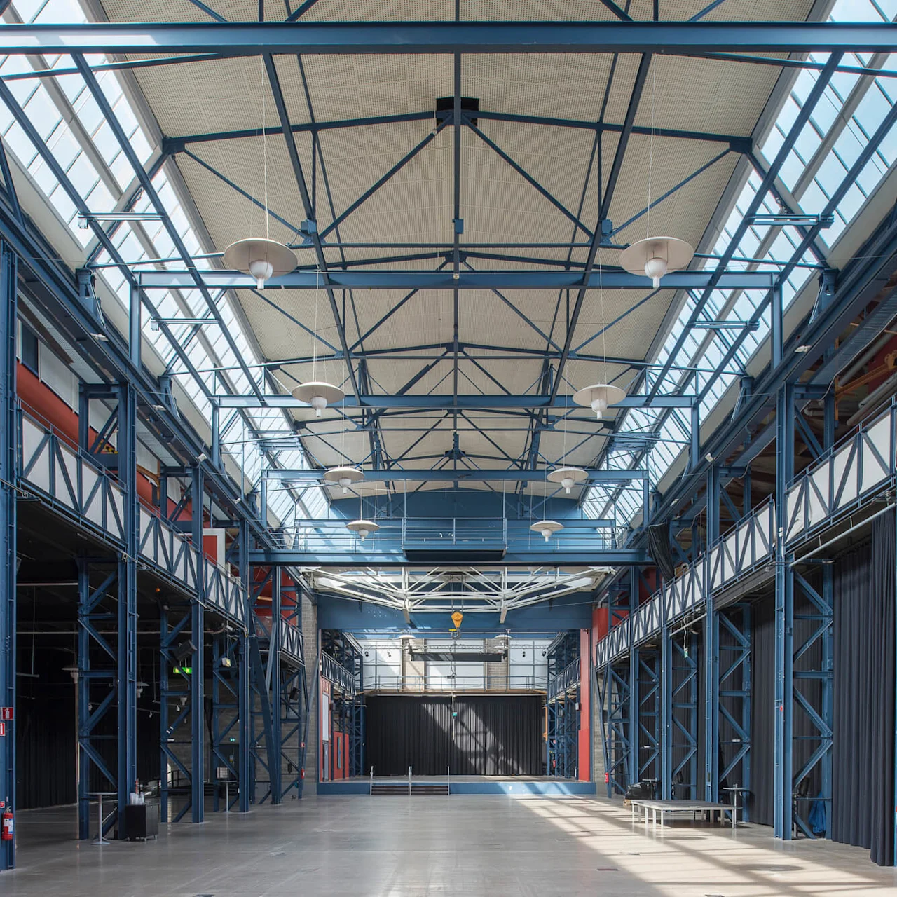 Spacious warehouse interior, featuring a high metal framework supporting a glass ceiling, with red and black walls on either side. Brightly lit through numerous skylights.