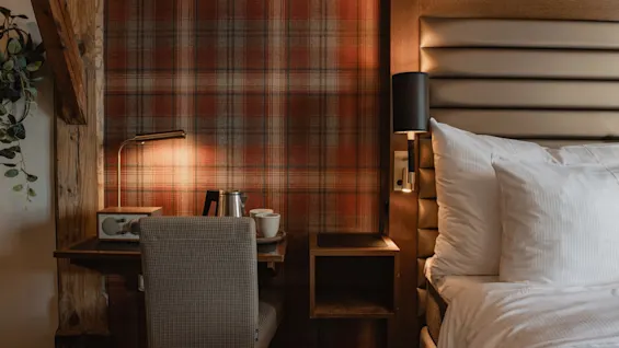 Et hyggeligt hotelværelse med en komfortabel seng, et lille skrivebord med en lampe og varmt skotskternet tapet, der skaber en indbydende atmosfære.