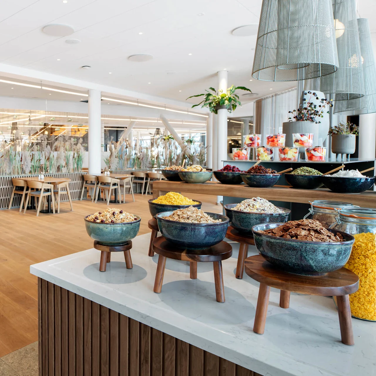 Boller med frokostblanding står på en benkeplate i et lyst, moderne spiseområde med tregulv og bord, omgitt av dekorative planter og hengende lys.