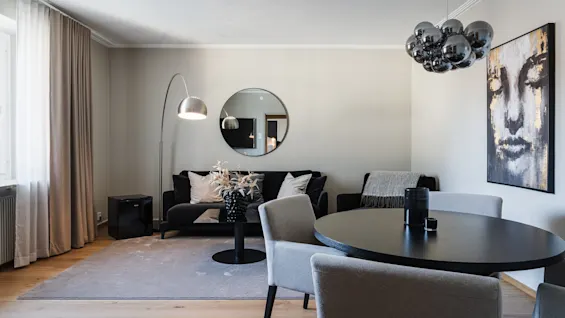 En svart soffa med vita kuddar står under en rund spegel; i närheten ett bord med grå stolar och en abstrakt ansiktsmålning. Ljusgrå väggar och trägolv fulländar rummet.