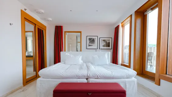A large bed with white linens is centered in a bright room. Wooden framed windows and doors surround, with red curtains and abstract art on the walls.