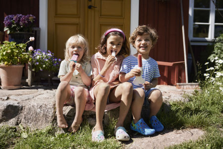 Tre barn sitter ute i solen på sommaren och äter glass i trädgården.