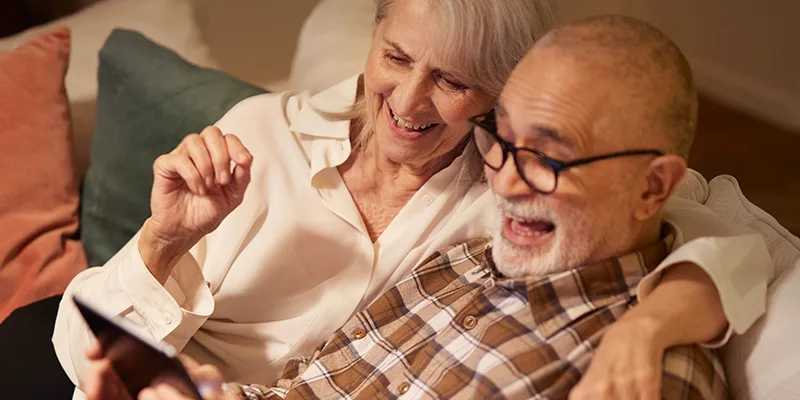 Ett äldre par håller om varandra i soffan och tittar på en surfplatta och skrattar tillsammans