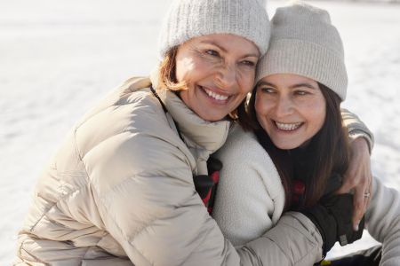 En äldre kvinna och hennes vuxna dotter sitter utomhus i solen på vintern, iklädda dunjackor och mössor.