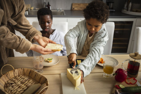 Familj sitter runt frukostbordet och äter tillsammans, ett barn skivar ost för att lägga på smörgåsen.