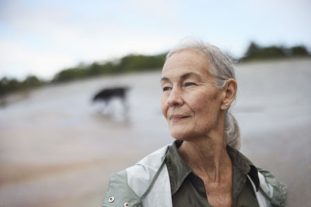 Äldre kvinna går runt ute på en strand med regnjacka.