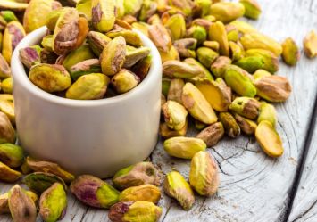 A white ceramic cup filled with unshelled pistachios, with additional pistachio nuts scattered on a rustic wooden surface. 