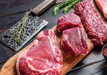 Fresh cuts of raw beef displayed on a wooden board with herbs nearby, illustrating the ongoing debate about whether dogs can eat raw beef or if raw food diets are beneficial, as many pet owners consider raw feeding for their dogs despite veterinary discussions about the safety of uncooked meat.