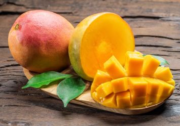 A vibrant, ripe mango shown both whole and cut in the distinctive cross-hatch pattern popular for dog-safe serving, displayed on a rustic wooden board with fresh green mango leaves,