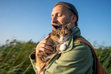 A man in a gray beanie and olive green jacket embraces his calico cat outdoors, eyes closed in a moment of pure contentment as tall grasses sway against a brilliant blue sky behind them.  This outdoor excursion captures the growing trend of people wondering can cats be service animals as they bring their feline companions into more aspects of daily life.