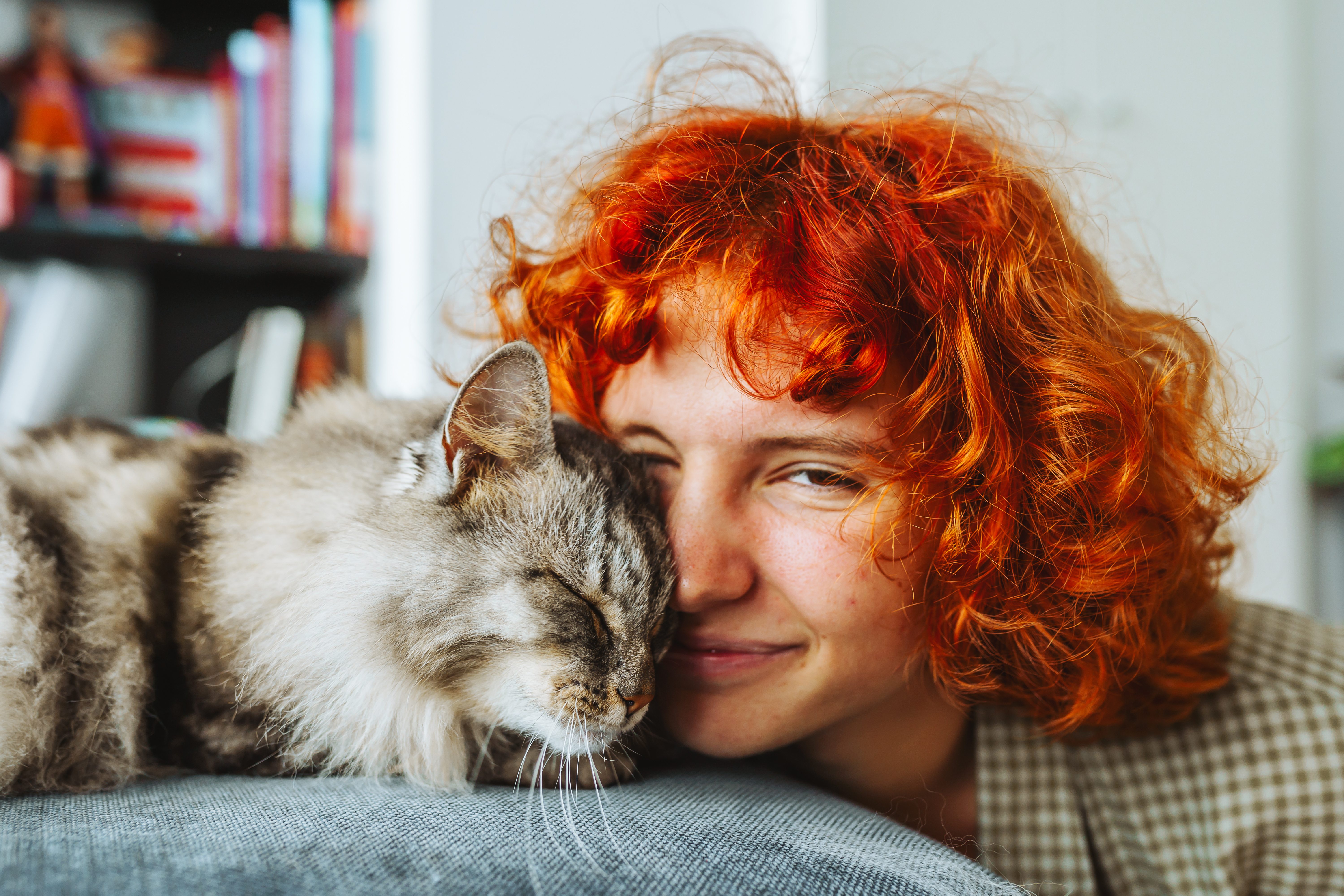 A young woman with curly red hair smiles contentedly as her gray tabby cat presses its face against hers on the couch — a classic velcro cat moment. If you've been wondering why your cat is so clingy, this kind of constant closeness is one of the most recognizable signs. 