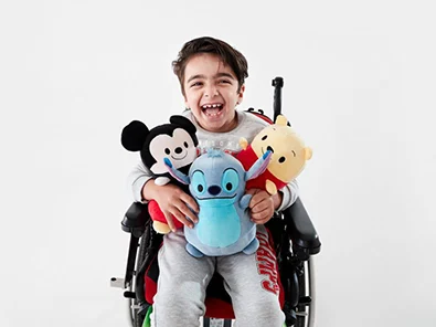 boy sitting in a wheelchair holding three disney plush toys 