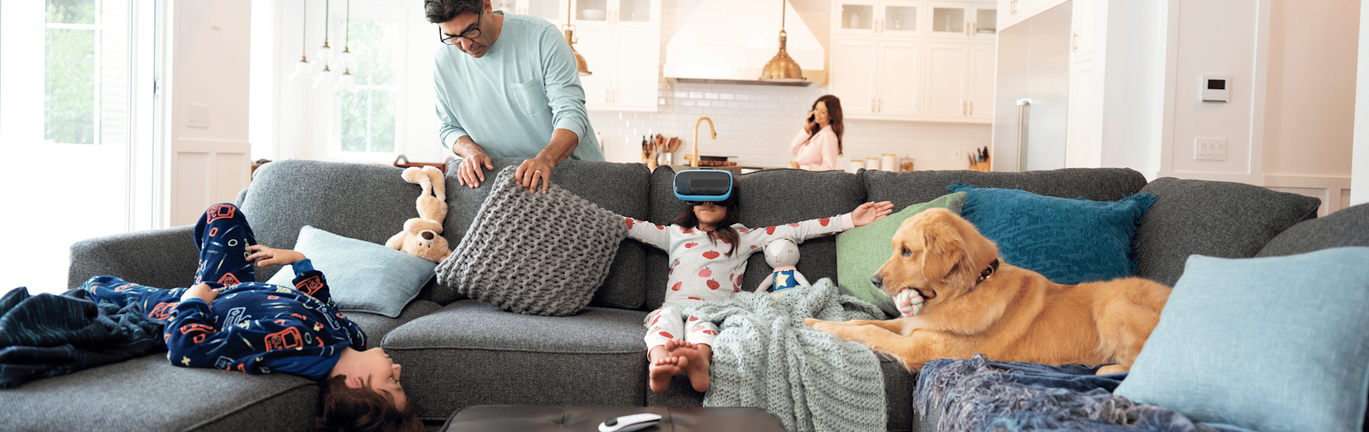 Man looking behind couch cushions with little girl sitting on couch wearing VR goggles next to a dog.