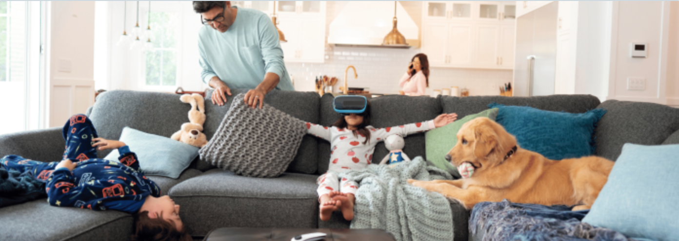 Man looking behind couch cushions with little girl sitting on couch wearing VR goggles next to a dog.