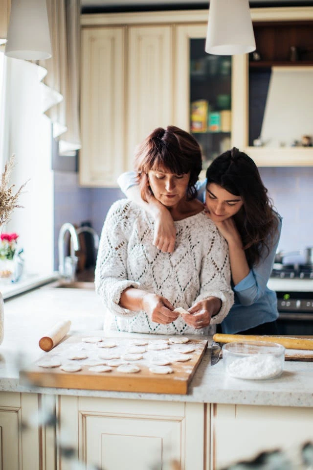 A teenage daughter has an arm around her mother in the kitchen while they make cookies together. 