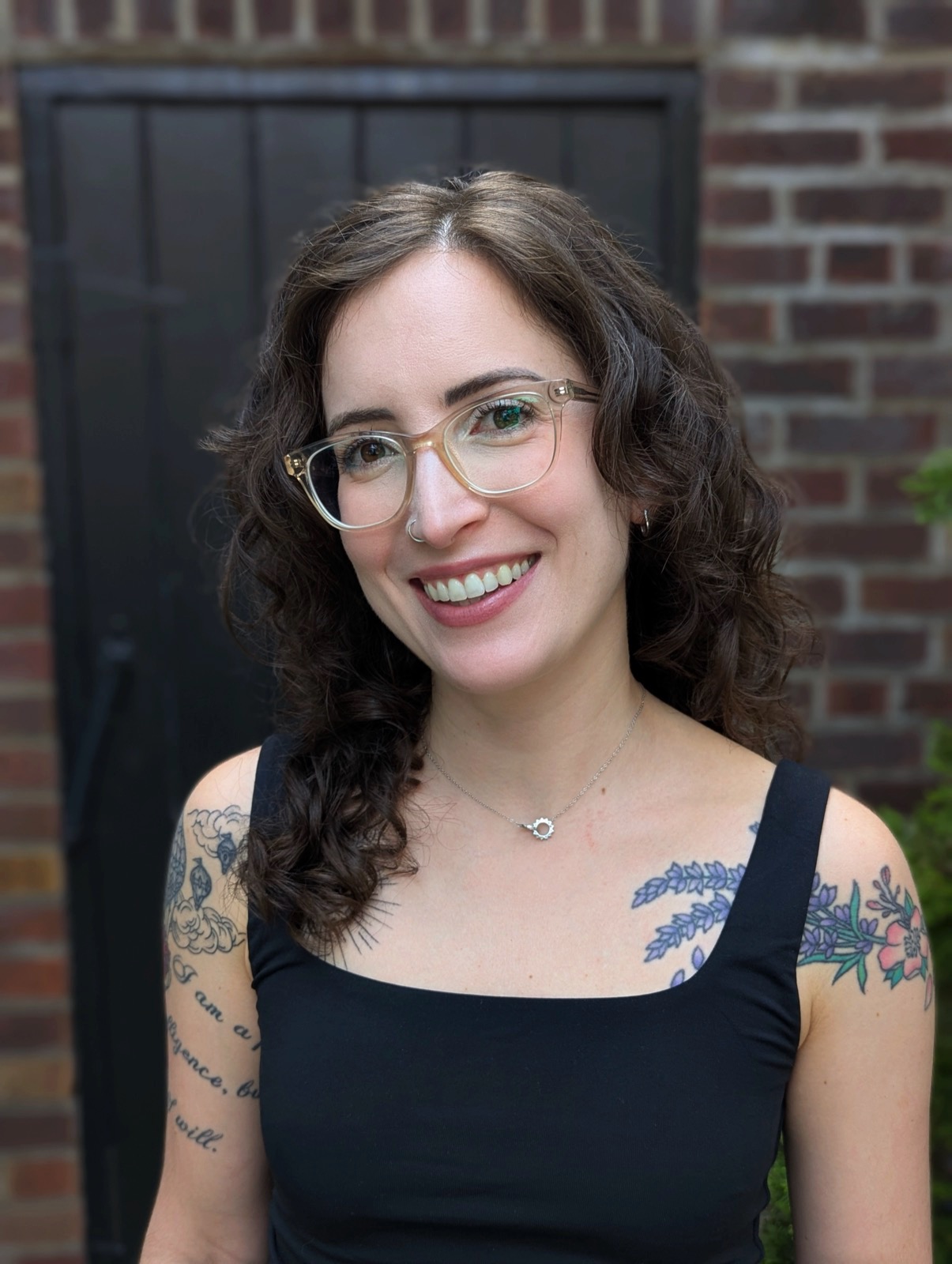 A headshot of Gianna Damis, owner of Created Self Therapy, wearing a black top and standing against dark background. She has upper body tattoos, clear frame glasses, and curly brown hair. 