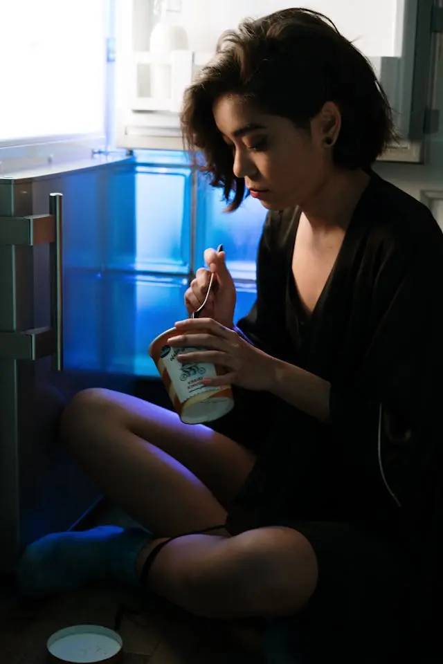 A sitting in front of the fridge, eating a pint of ice cream and looking sad.