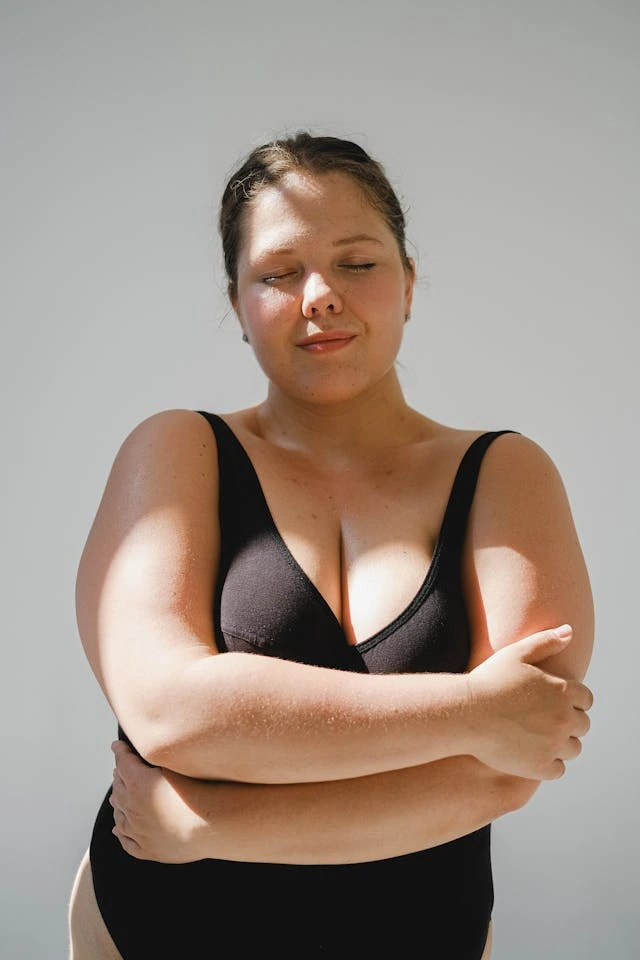 A plus size woman stands with her eyes closed and the sun on her face. She hugs her body and smiles softly. 
