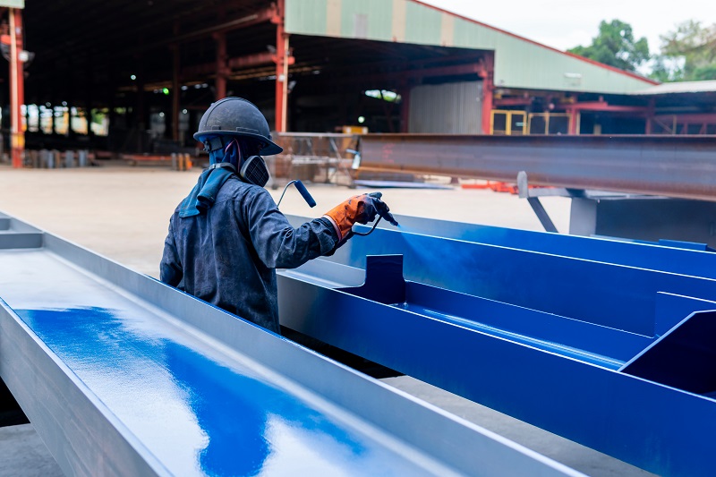 A man wearing a helmet and protective gear spraying blue paint on a metal beam.