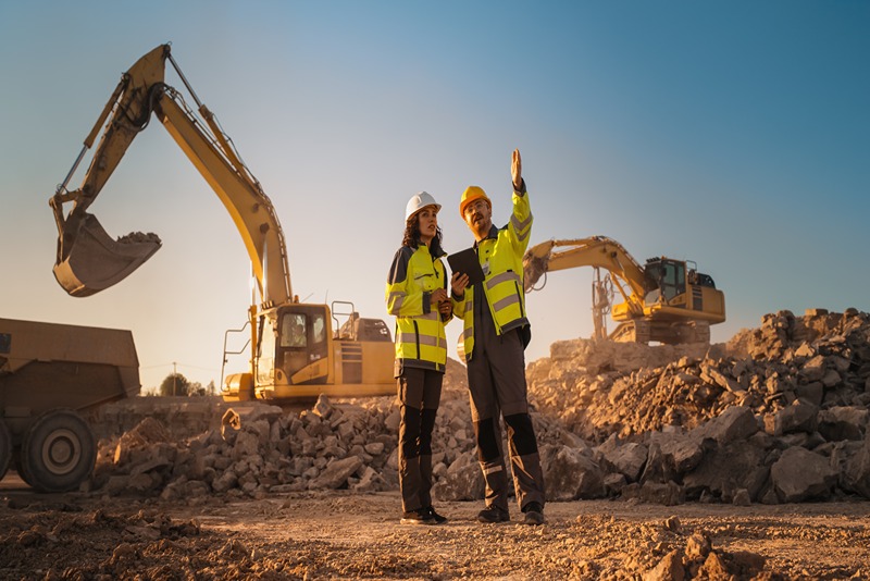 Hispanic_Female_Inspector_Talking_to_Caucasian_Male_Land_Development_Manager_With_Tablet_On_Construction_Site_Of_Real_Estate_Project._Excavators_Preparing_For_Laying_Building_Foundation._Hot_Sunny_Day