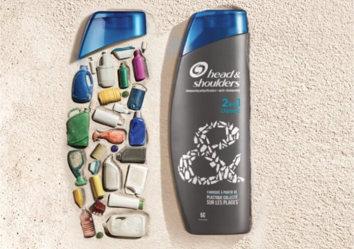 Twee shampooflessen met achtergrond strand. Beiden dezelfde blauwe dop. Linker fles gevormd uit plastic flessen. Rechterfles is grijs. 2 en 1 classic.