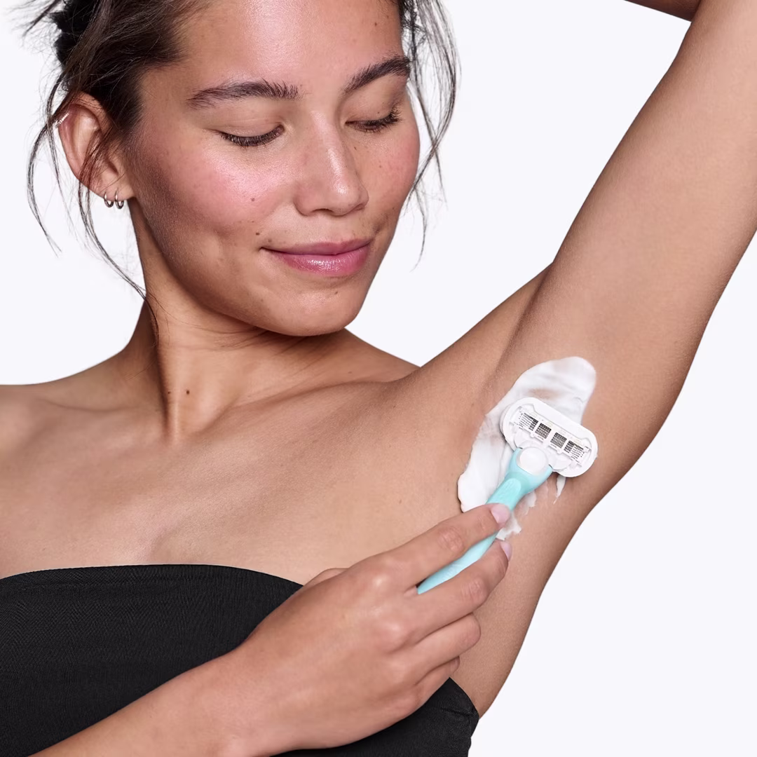 Žena v černém topu si holí levé podpaží modrým holicím strojkem. Žena v černém topu si holí levé podpaží modrým holicím strojkem.