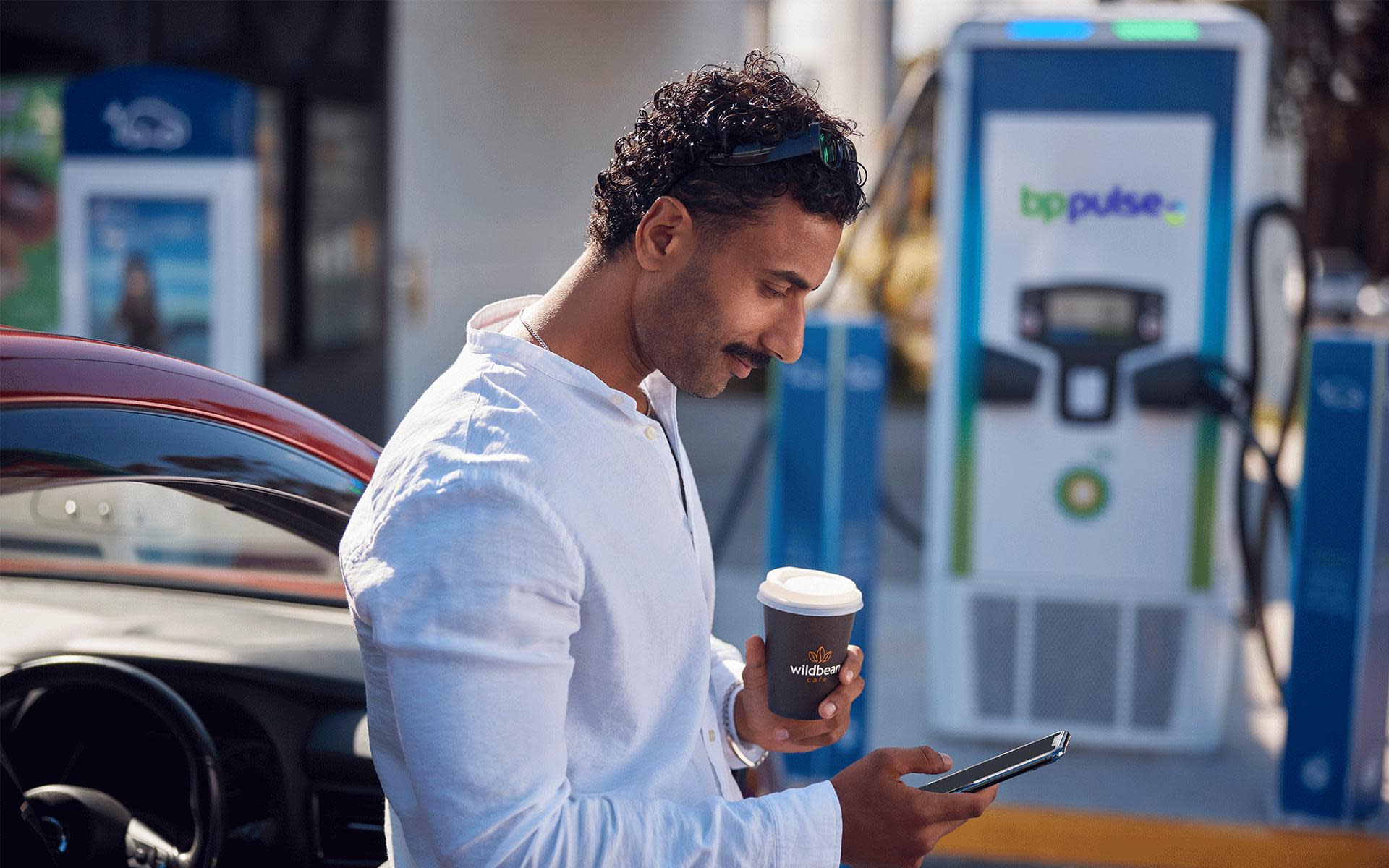 A man holds his phone and a drink from Wild Bean as his electric vehicle charges on the bp pulse network.